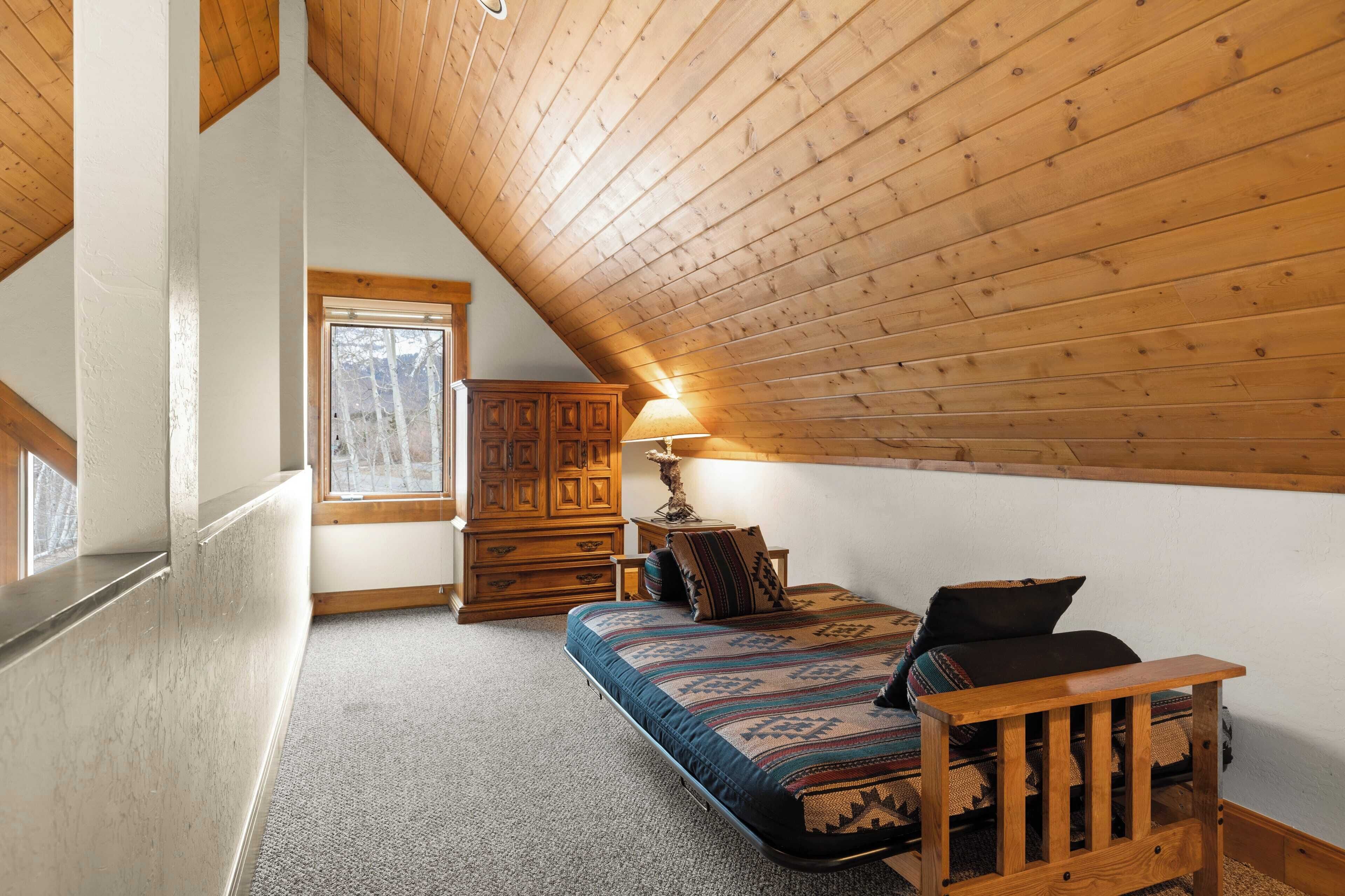 Ein Schlafzimmer im Loftsstil mit schrägem Dach, einem Bett mit gemusterten Betttextilien, einem Holznachttisch, einer Kommode und einem Fenster mit Blick auf Bäume.
