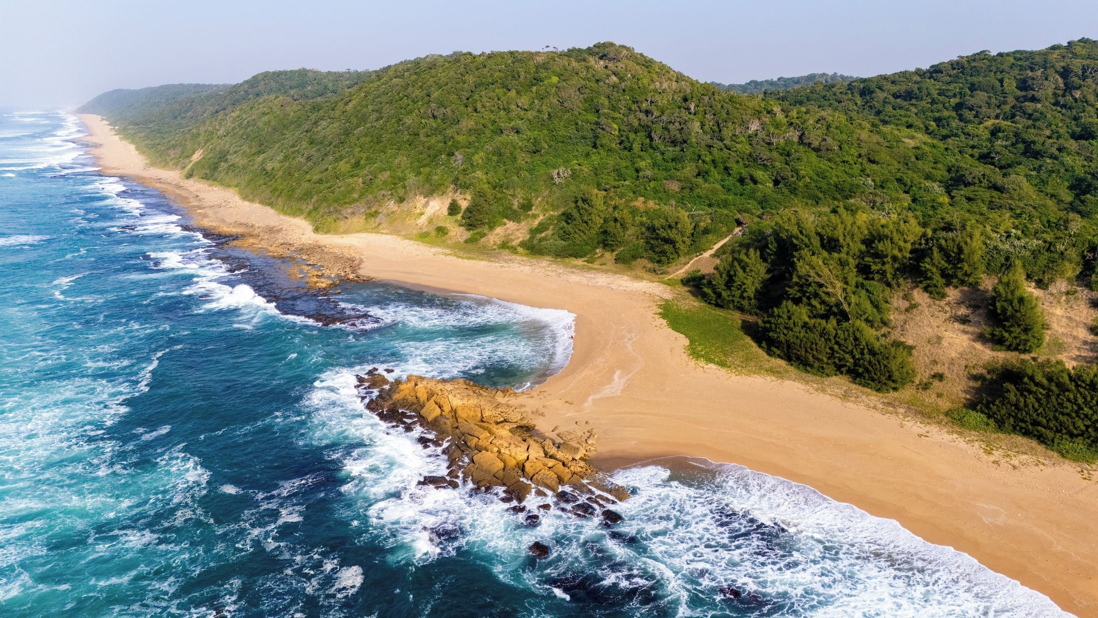 Eine Küstenlandschaft mit Sandstrand, Felsen und dicht bewachsenen grünen Hügeln.