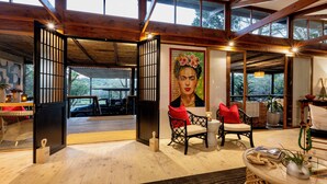 A spacious room with wooden floors, a large window, and a view of the outdoors. There is a painting on the wall, a round table with a plant, and a chair with a red cushion. - Makakatana Bay Lodge (Makakatana)
