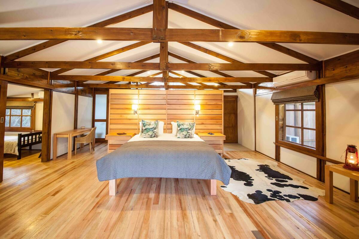 A bedroom with a wooden ceiling, a bed with a grey bedspread, two bedside lamps, a cowhide rug, and a wooden bench.