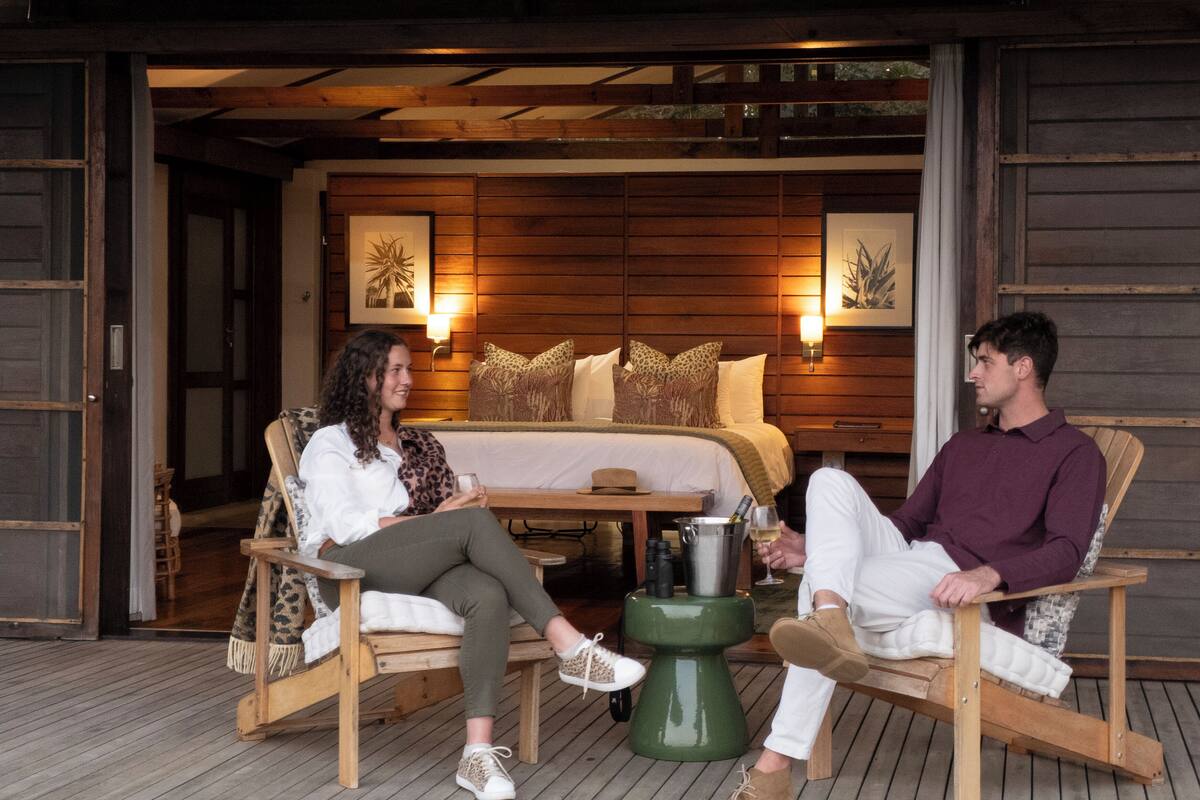Two people seated on wooden chairs on a wooden deck, with a bed and wall-mounted lights in the background.