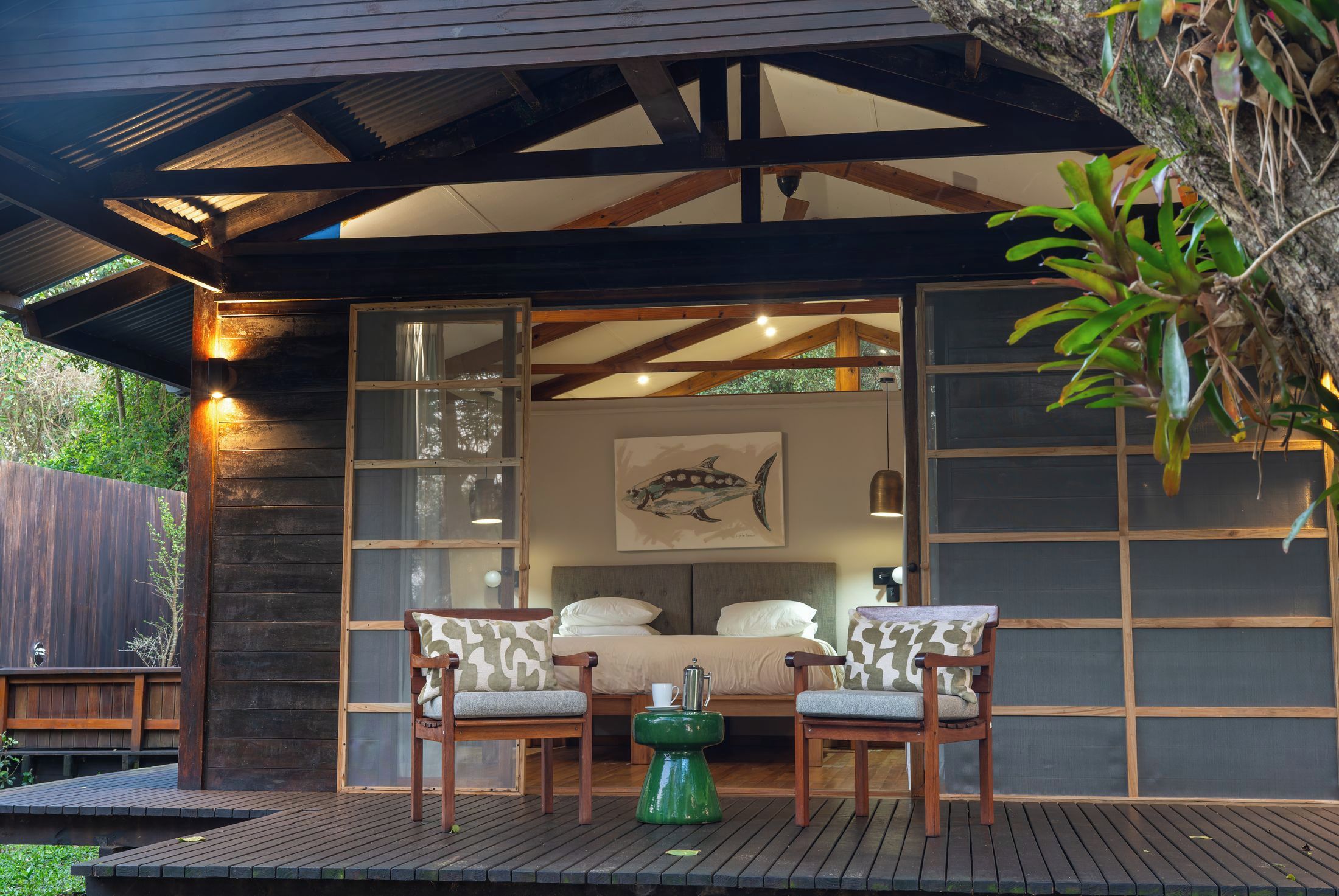A cozy outdoor seating area with a wooden bench and a table, a bed with a headboard, and a framed fish painting on the wall.
