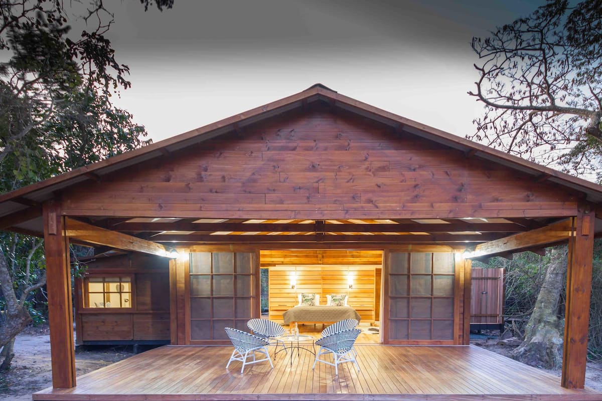A wooden cabin with a covered porch, a bed, and two chairs.