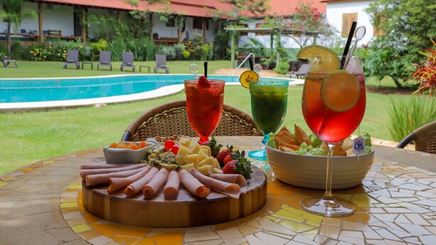 A table with a charcuterie board, two drinks, and a bowl of chips and dip, set outdoors with a pool and lounge chairs in the background. - Hotel Fazenda Bela Vista (Dourado)