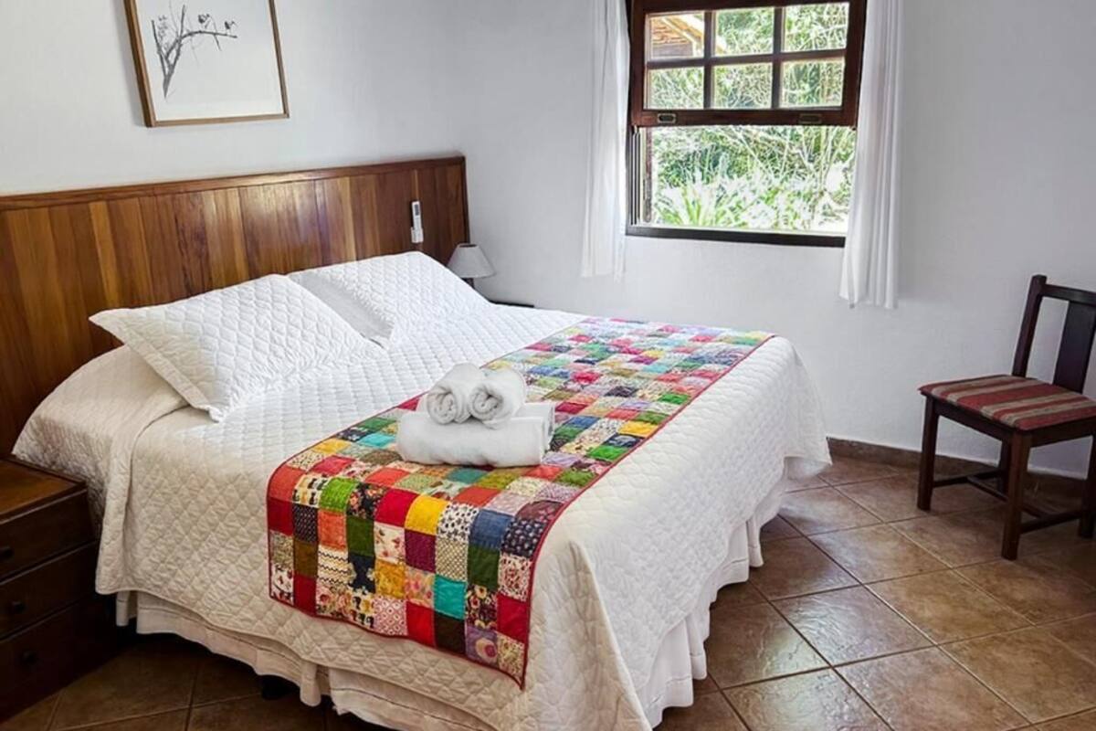 A neatly made bed with a quilt, a wooden headboard, a window with a view, a chair, and a nightstand.