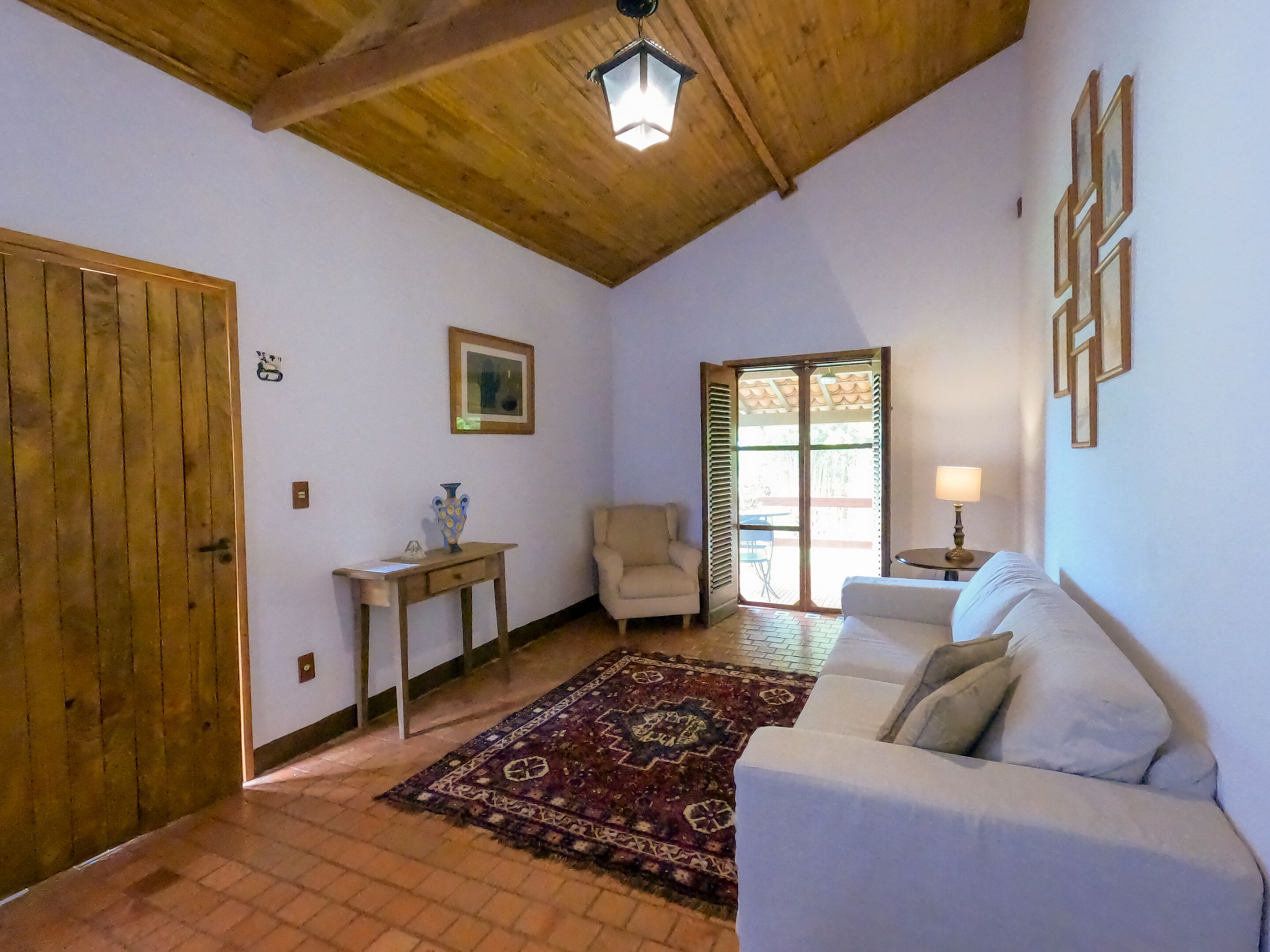 A cozy living room with a wooden ceiling, a white sofa, a patterned rug, a small table with a vase, and a chair.