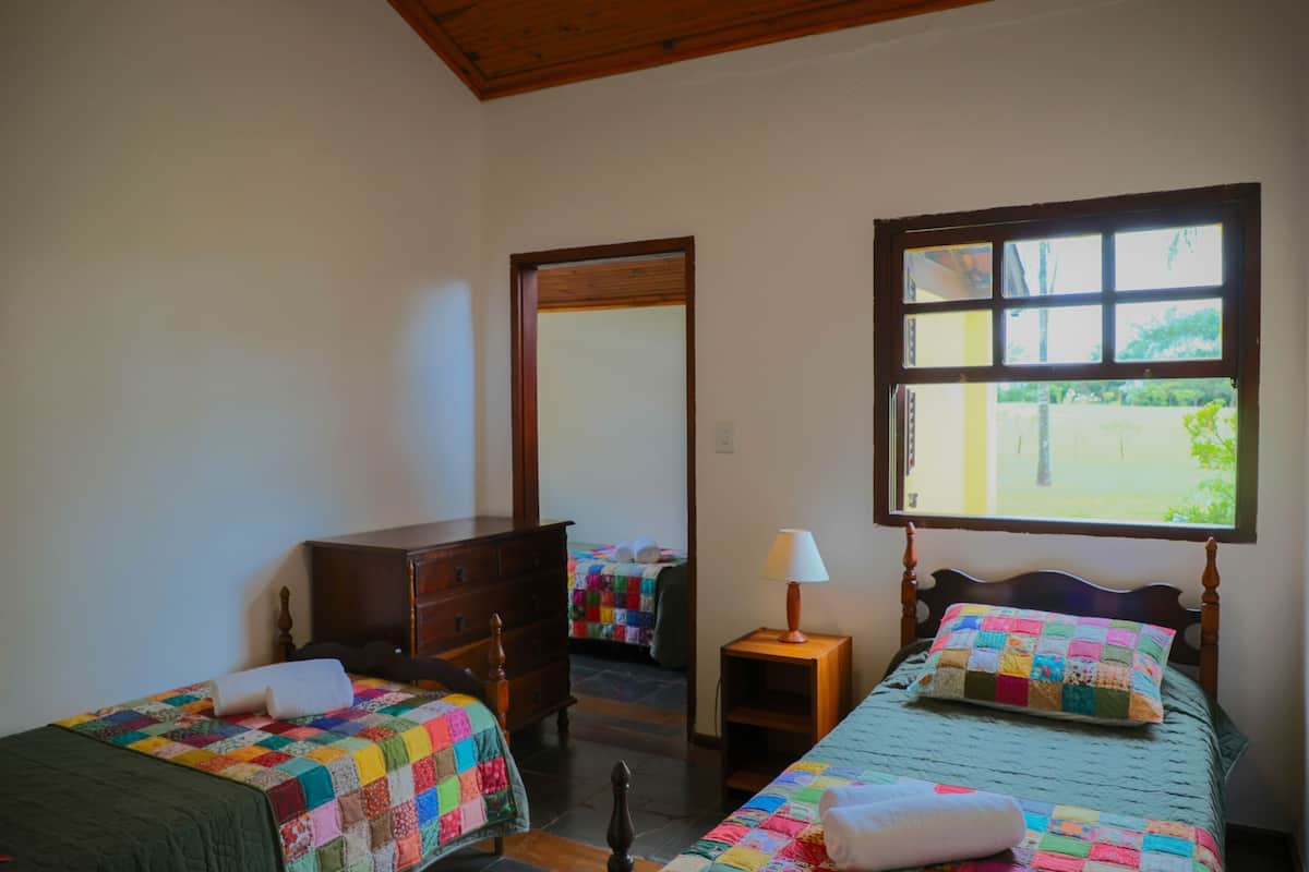 A bedroom with two beds, a dresser, a nightstand with a lamp, and a window with a view of greenery.
