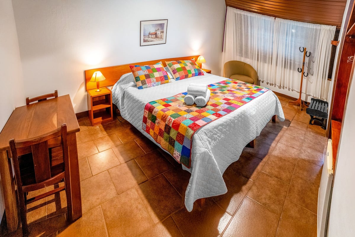 A neatly arranged hotel room with a bed, a wooden nightstand with a lamp, a wooden chair, and a small table.