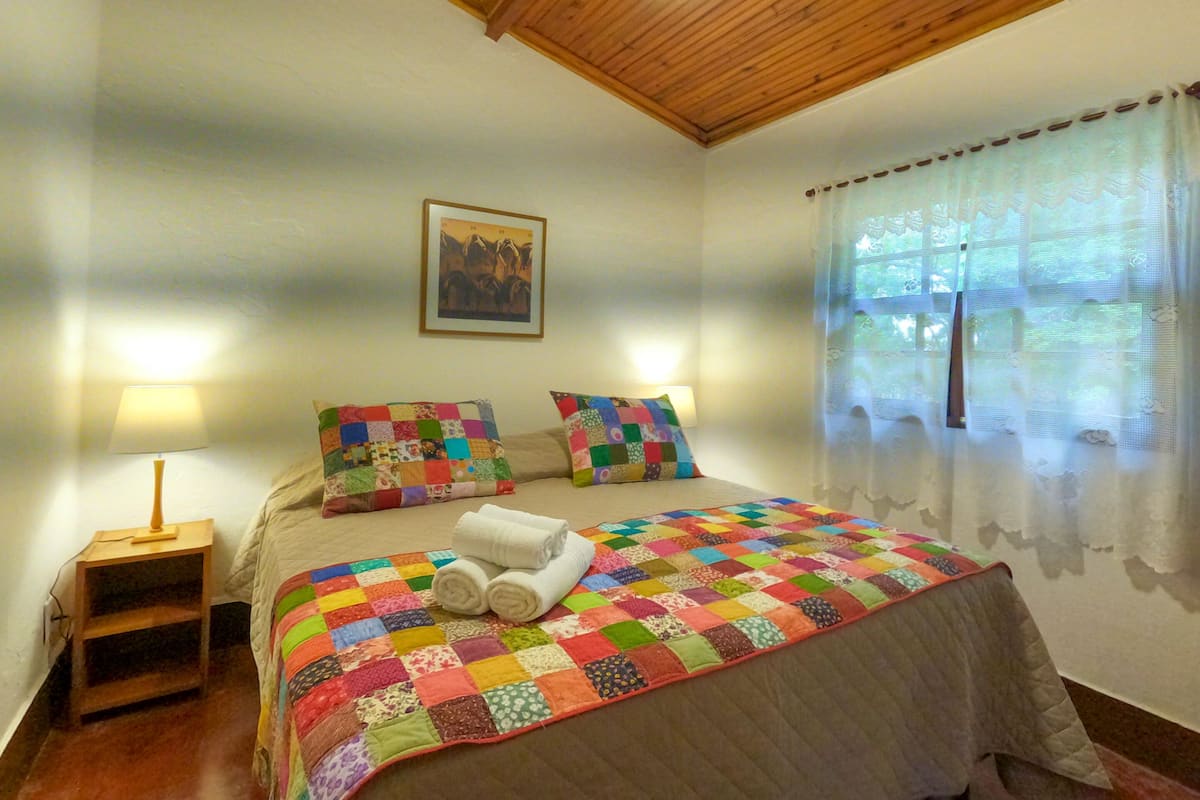 A bedroom with a wooden ceiling, a quilted bedspread, two pillows, a bedside table with a lamp, and a window with sheer curtains.