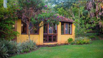 Un petit bâtiment jaune, surmonté d’un toit en tuiles, entouré d’une végétation luxuriante et de plantes en fleurs.
