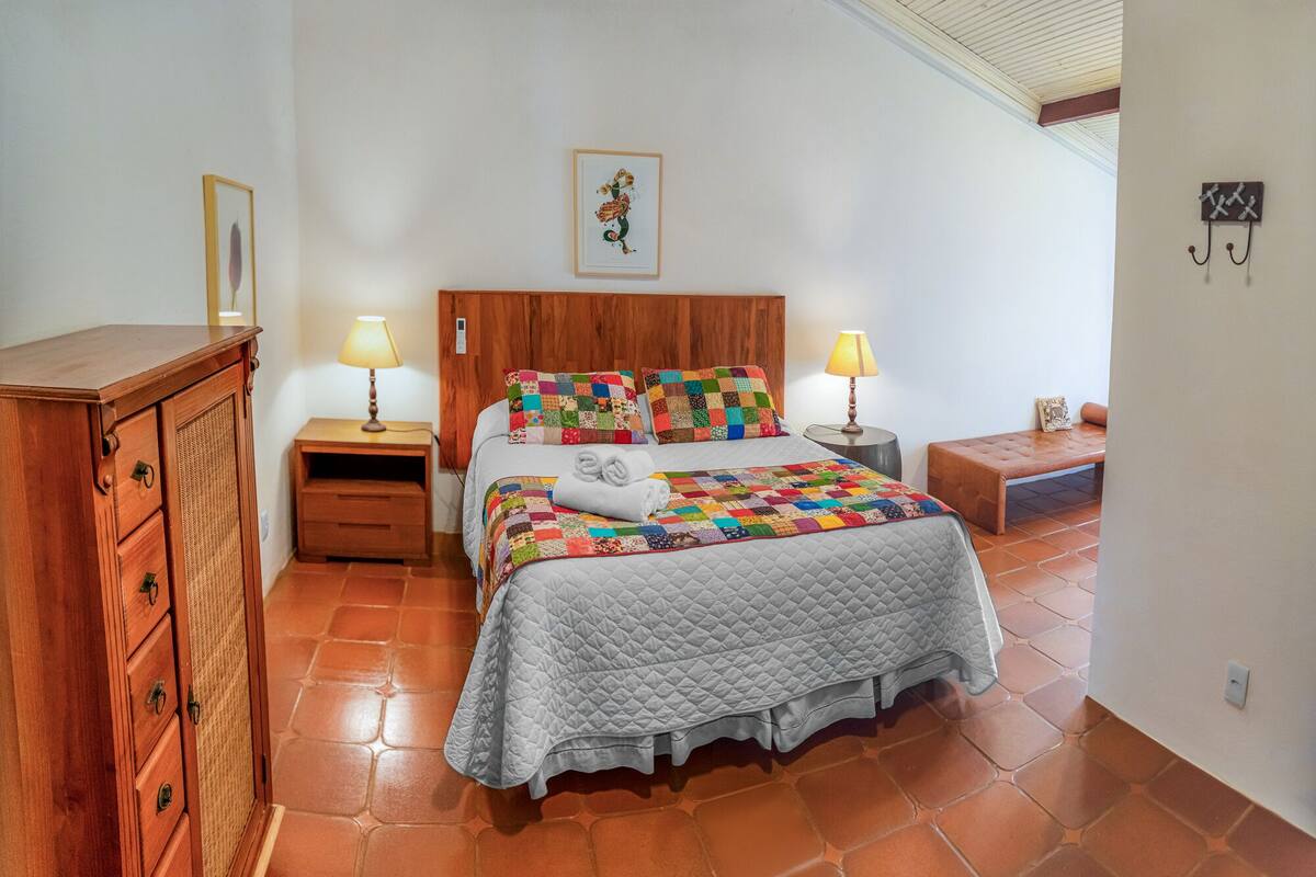 A bedroom with a wooden headboard, a quilted bedspread, two bedside lamps, a wooden nightstand, a bench, and a wall-mounted coat rack.