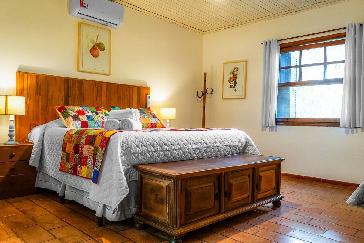 A bedroom with a wooden headboard, a quilted bedspread, a wooden nightstand with a lamp, a wooden bench, and a window with curtains.
