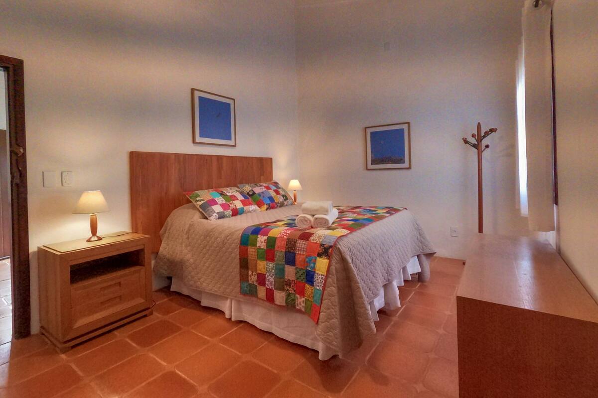 A bedroom with a wooden headboard, a quilted bedspread, a bedside table with a lamp, and a wooden coat rack.