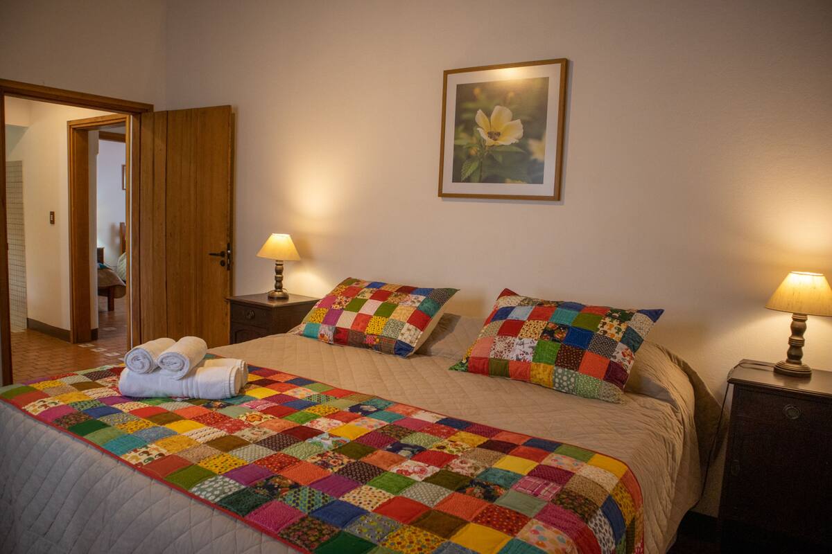 A neatly made bed with a colorful quilt, two pillows, and a rolled towel on the bed. There are two bedside tables with lamps, a framed picture on the wall, and a door leading to another room.