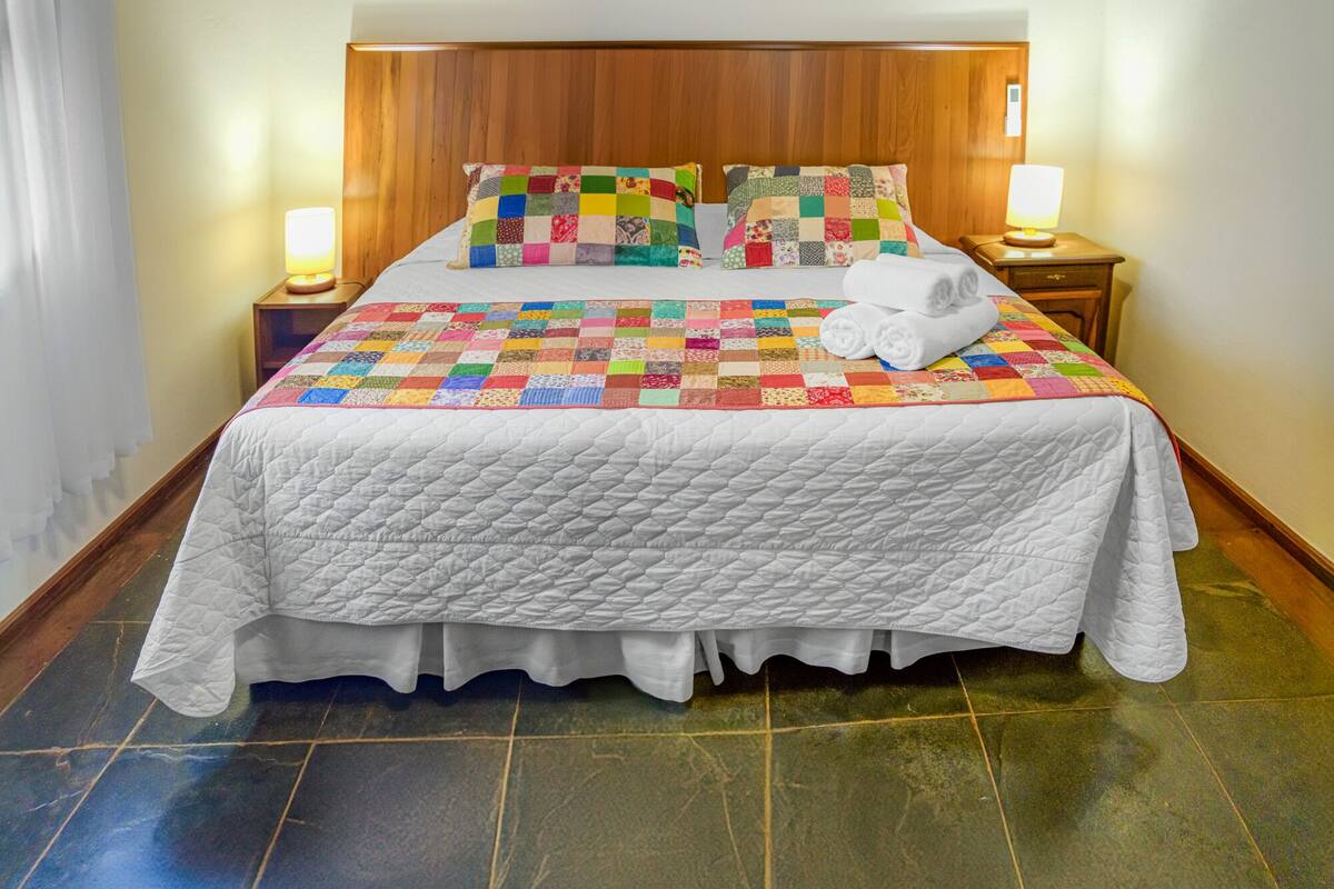 A neatly made bed with a quilt, two pillows, and a rolled towel on a wooden headboard. There are two bedside lamps on wooden nightstands.