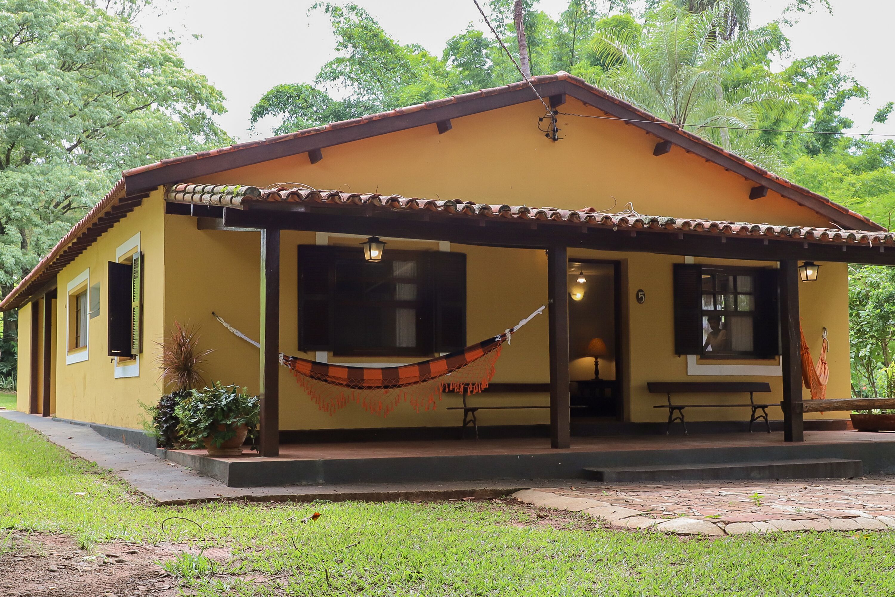 A yellow building with a tiled roof, a porch with a hammock, and a bench.