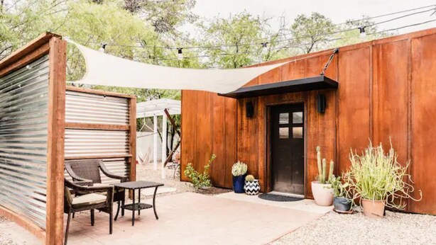 Un bâtiment rustique en bois, doté d’une porte, d’un patio couvert avec des chaises et de plantes en pot.