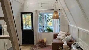 Una camera da letto mansardata accogliente, con soffitto spiovente, una ventola a soffitto, una finestra con tende, un divano marrone con cuscini, un tappeto piccolo e un tavolo di legno con un portaconfetti.