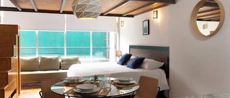 A hotel room with a bed, a dining table with chairs, a large window, and a ceiling light fixture.