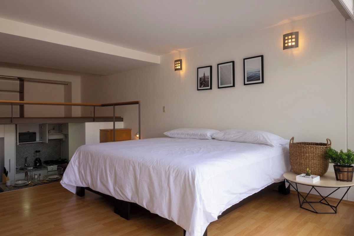 Een slaapkamer met een groot bed, een klein tafeltje met een plant en een keukenruimte zichtbaar door een open deur.
