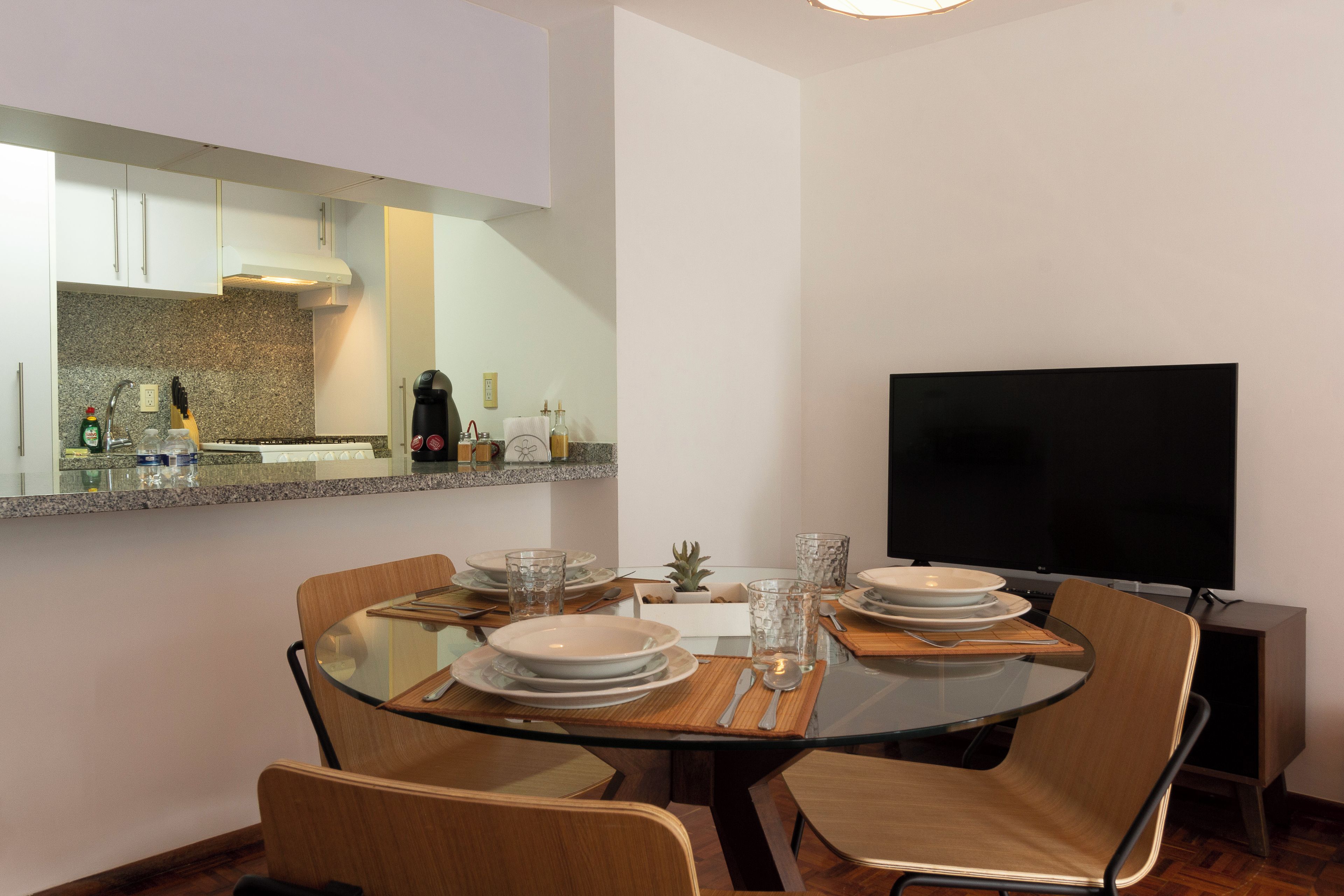 Uma área de refeições moderna com mesa de vidro, cadeiras de madeira e uma TV de tela plana.