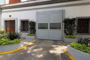 Une entrée fermée par un portail et dotée d’une double porte, une petite fenêtre ornée d’une grille métallique et un jardinier avec des plantes vertes.