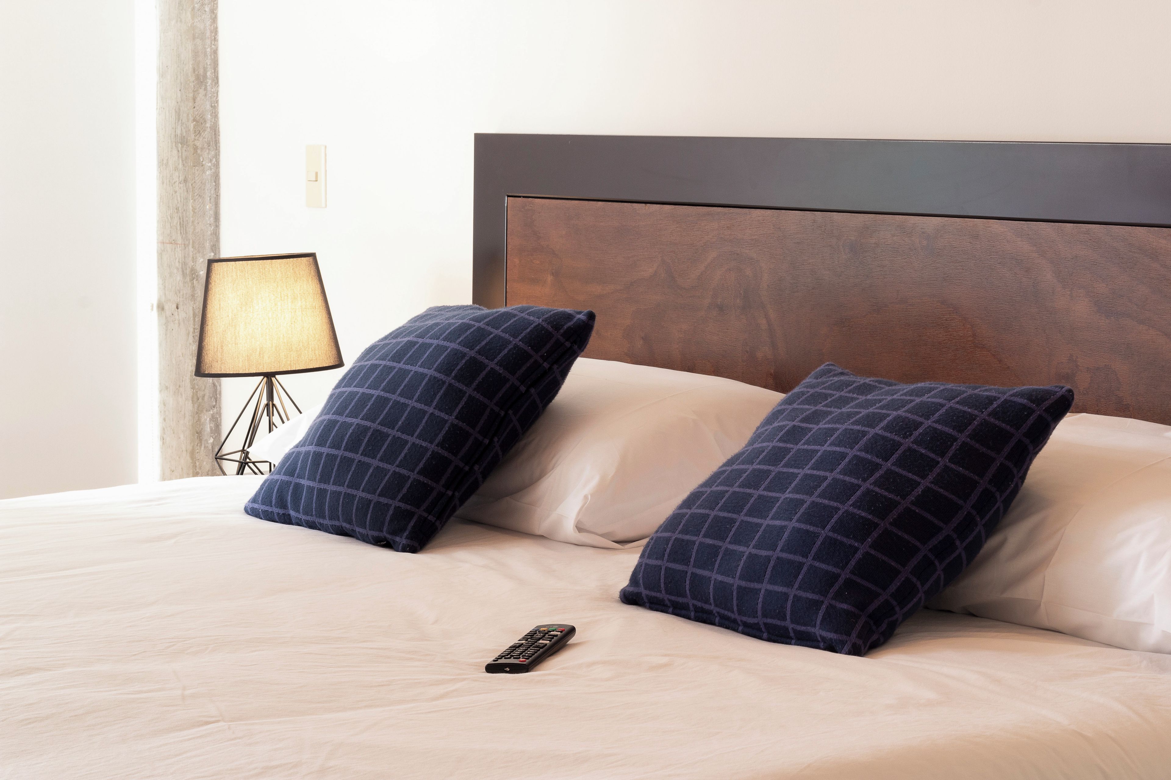 Una cama bien hecha con dos almohadas oscuras a cuadros, un control remoto sobre la cama y una lámpara en una mesita de noche.