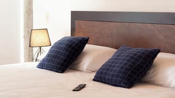 Una cama bien hecha con dos almohadas oscuras a cuadros, un control remoto sobre la cama y una lámpara en una mesita de noche.