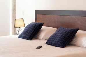 Un lit bien fait, orné de deux coussins à carreaux foncés, une télécommande posée sur le lit et une lampe sur une table de chevet.