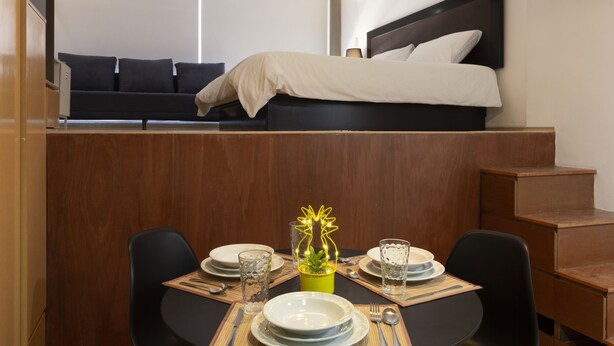 A compact hotel room with a bed, a small dining area with a table set for two, and a sofa in the background. - Prosperidad By HomiRent (Mexico City)