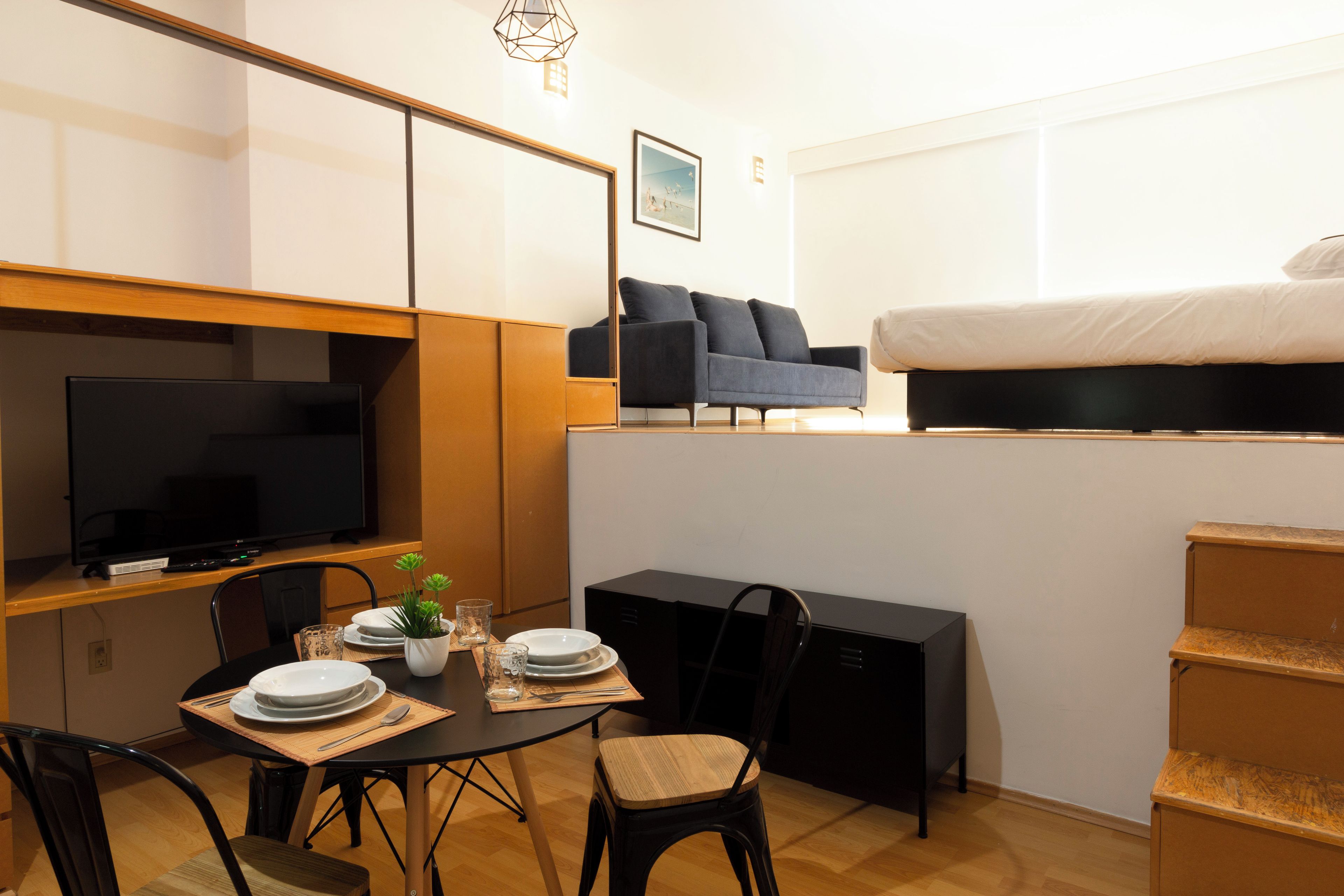 Habitación de hotel pequeña con cama, televisión, mesa de comedor con sillas y una kitchenette.