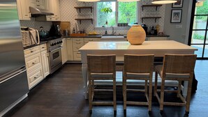 Een moderne keuken met witte kasten, een groot eiland met barkrukken, hanglampen en uitzicht op groen buiten.