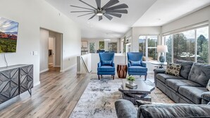 A spacious living room with a modern ceiling fan, a large window offering a view of the outdoors, and a cozy seating area with a sofa and armchairs. - Experience luxury in this spacious 4-bedroom Sedona Maxwell Ridge with stunning red rock views. (Sedona)