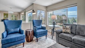 A spacious living room with two blue armchairs, a grey sofa, a round wooden side table, and a floor lamp. Large windows provide a view of the outdoors. - Experience luxury in this spacious 4-bedroom Sedona Maxwell Ridge with stunning red rock views. (Sedona)