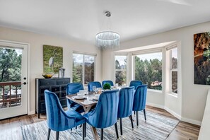 Uma sala de jantar com mesa de madeira, cadeiras estofadas em azul, um candelabro e vista para árvores através de grandes janelas.