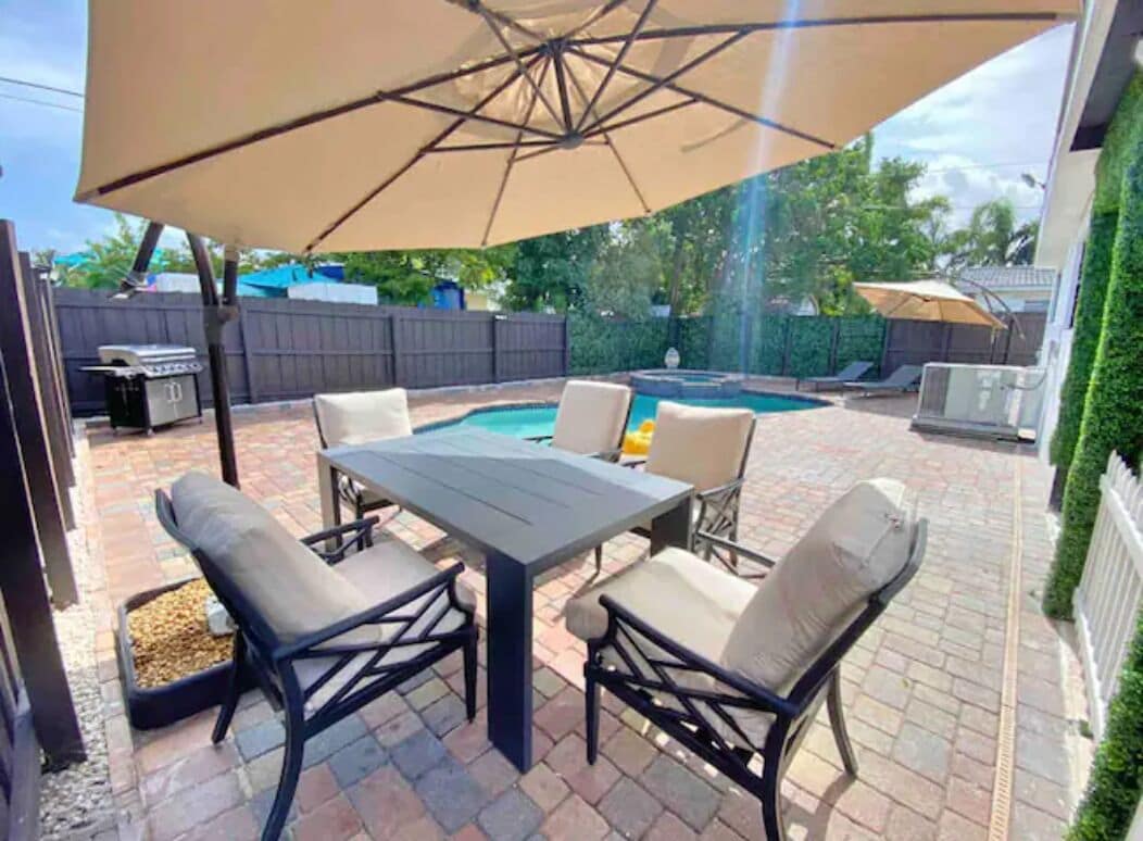 En udendørs terrasse med borde og stole, en pool og et overdækket område med en parasol.