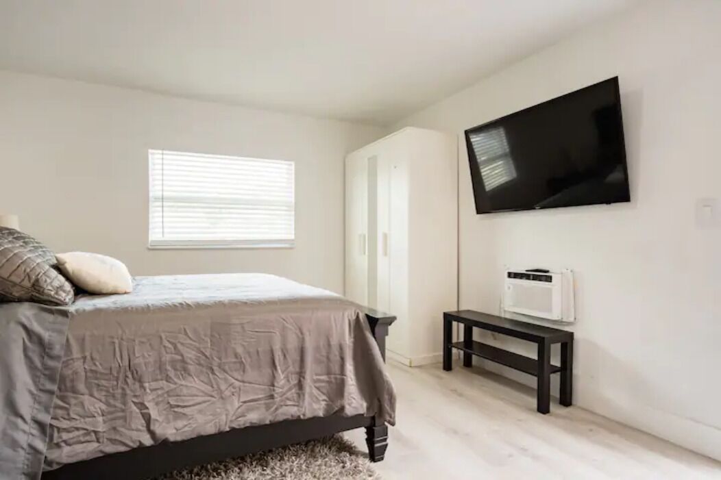 A bedroom with a bed, a TV mounted on the wall, a heater on a small table, and a window with blinds.
