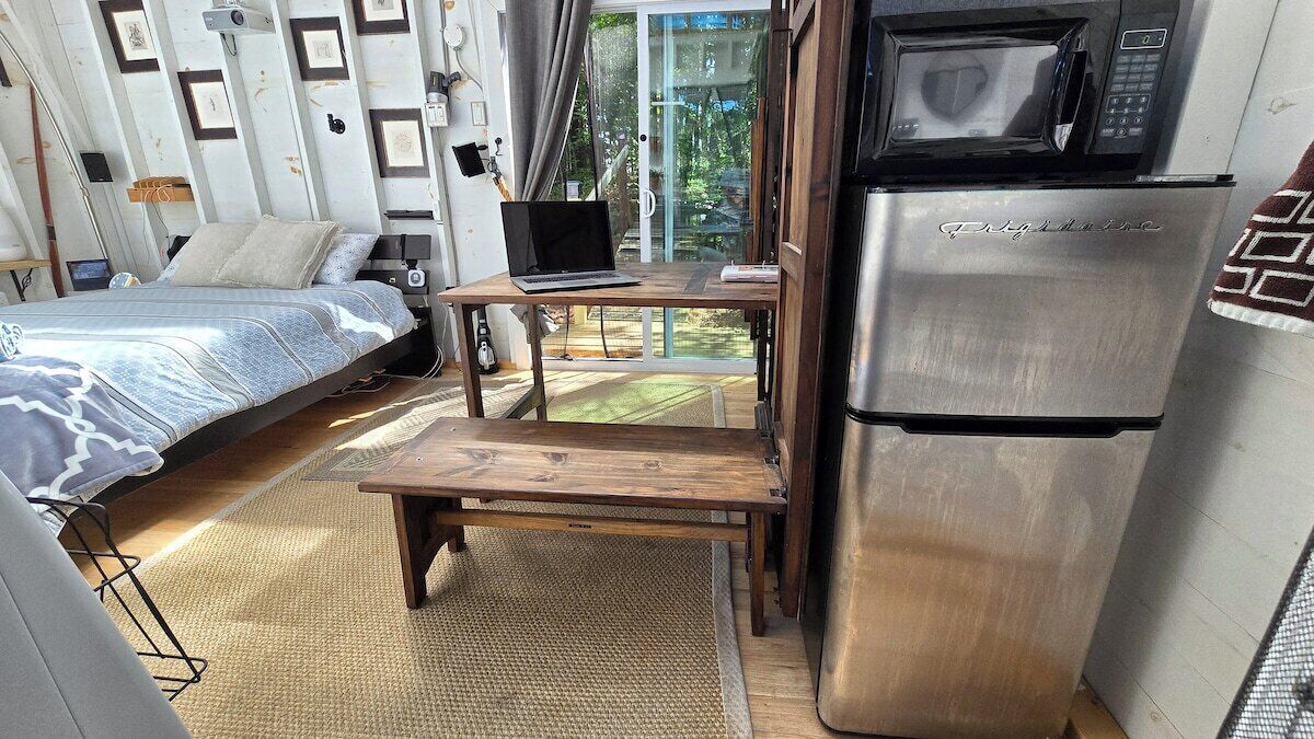 Une pièce compacte comprenant un lit, un ordinateur portable posé sur une petite table, un réfrigérateur et une porte-fenêtre coulissante donnant sur un balcon.