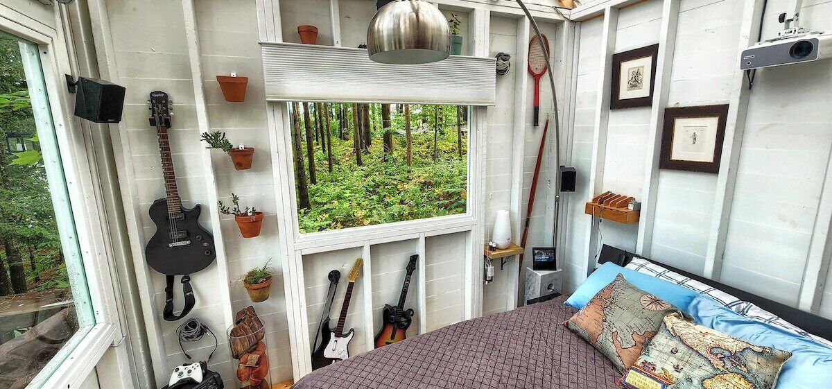 Ein Schlafzimmer mit einem Bett, Gitarren, Topfpflanzen und einem Fenster mit Blick auf einen Wald.