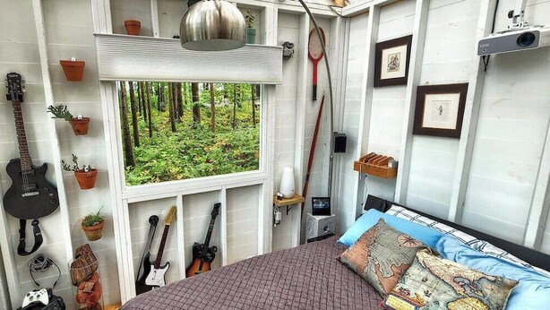 A bedroom with a bed, guitars, potted plants, and a window with a forest view. - Stabbin Cabin #2 on Grant Island-HotTub,Boat,Pets (BRANTINGHAM)