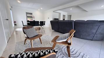 A spacious living room with a grey sectional sofa, two wooden chairs with patterned cushions, a dining table with chairs, and a dog.
