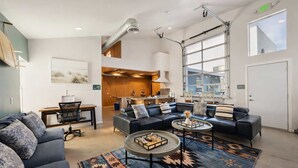 A spacious living area with a large black leather sofa, a round coffee table, and a patterned rug. There is a kitchen area visible in the background with wooden cabinets and a large window. - Resort Perks: 17-bed Surf lof.t private deck (San Diego)