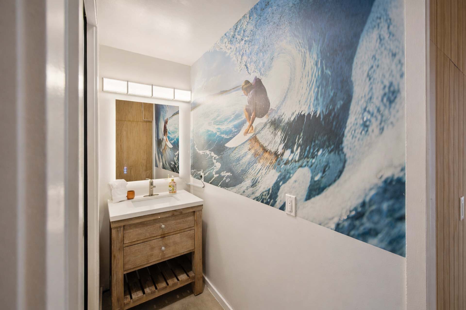 Een badkamer met een houten wastafelmeubel, een grote spiegel en een muurschildering van een surfer die een golf pakt.