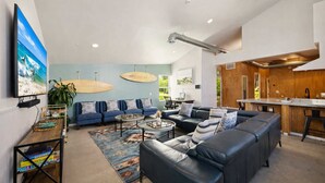 A modern living room with a large flat-screen TV, a sofa set, a coffee table, and a rug. There are surfboards on the wall and a kitchen area with wooden cabinets. - Resort Perks: 17-bed Surf lof.t private deck (San Diego)