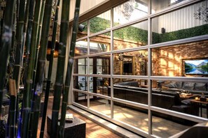 Un restaurante con un amplio ventanal, plantas de bambú y paredes interiores de ladrillo.