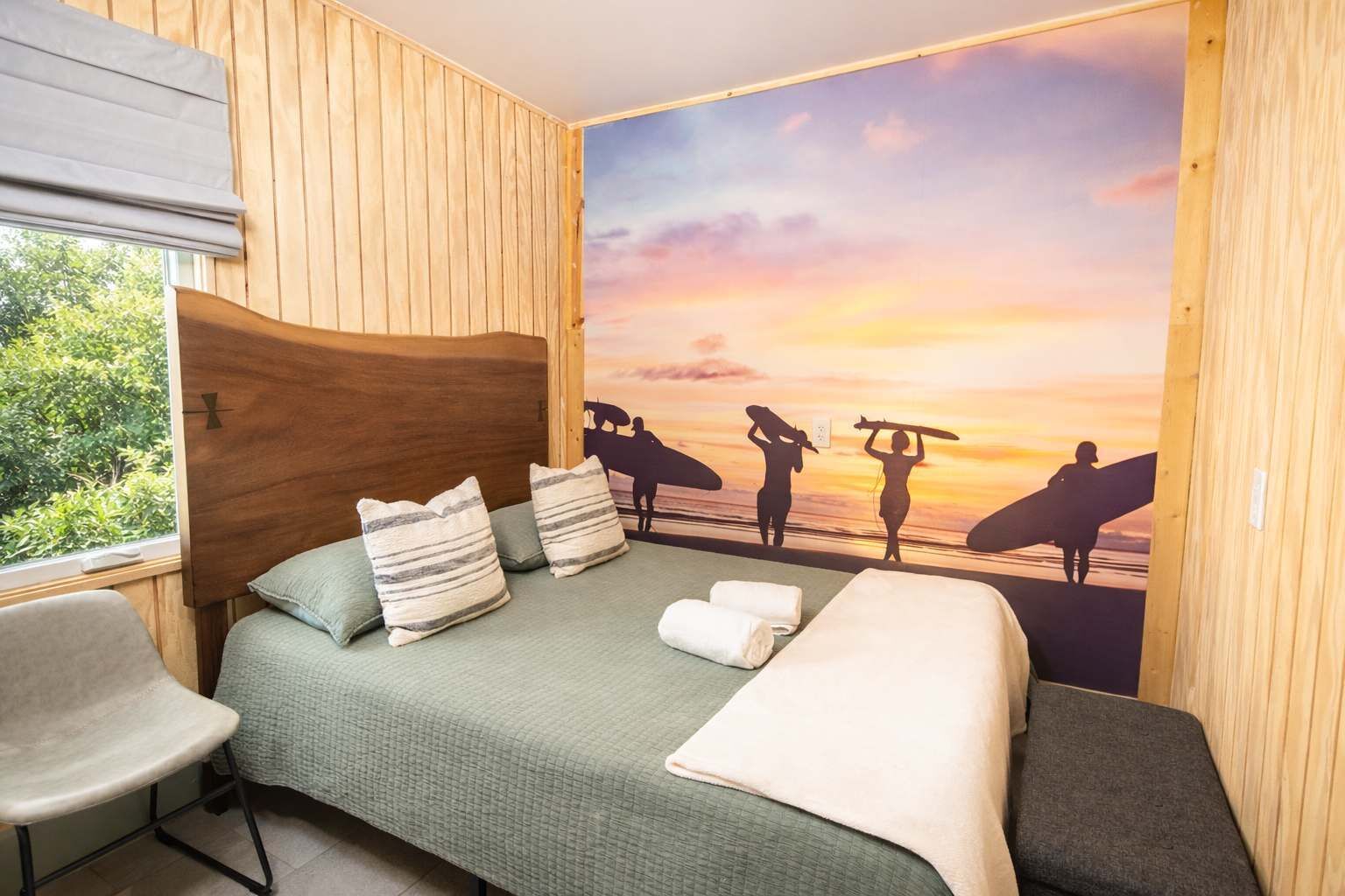 A room with a bed, a chair, and a wall mural of surfers at sunset.