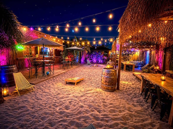 Eine Strandbar mit Außenbereich, Sitzgelegenheiten, Lichterketten und einem Strohdachhaus.