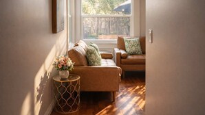A cozy living room with a brown sofa, a side table with a vase of flowers, and a window with a view of greenery. - Bamboo House  (Downtown, Convention Center) 3BR (San Diego)