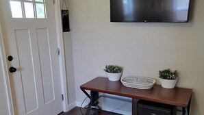 A living room with a flat-screen TV mounted on the wall, a wooden coffee table with two potted plants, a printer, and a door with a window. - Cozy 3/2 cottage near Memorial Hospital. (Lake Charles)