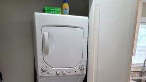A compact laundry room with a front-loading washer and dryer, a mop, a broom, and cleaning supplies. - Cozy 3/2 cottage near Memorial Hospital. (Lake Charles)